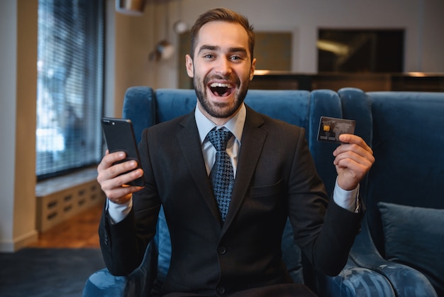 Handsome young businessman wearing suit sitting at the hotel lobby, using mobile phone, showing plastic credit card