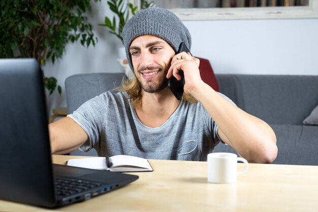 Фото Красивый молодой кавказский человек с помощью компьютера работает дома и разговаривает с мобильным телефоном, чувствуя себя счастливым