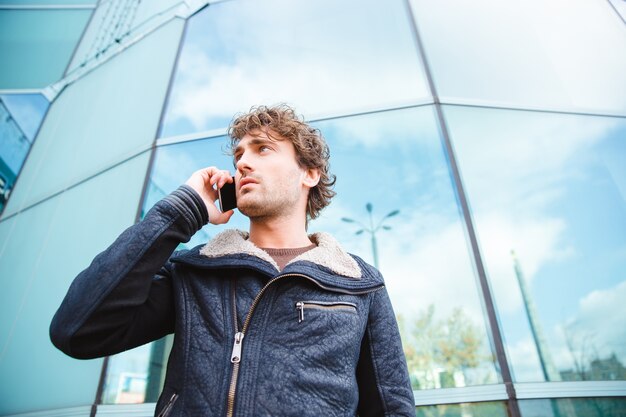 Handsome young confident attractive successful curly guy in black jacket talking on cellphone near modern glass building