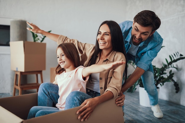 Handsome young dad pushing a cardboard box with his wife and child inside while playing in the new house