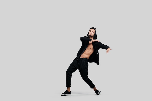 Handsome young dancer of street dancing dressed in black pants, a sweatshirt on a naked torso and a hood dances on a white