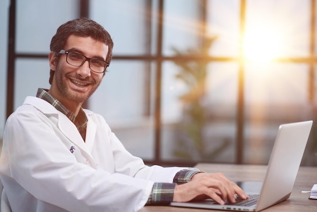 A handsome young doctor sits at a table and smiles friendly while looking at the camera