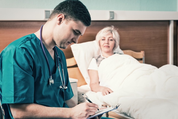 Handsome young doctor in uniform with phonendoscope on his neck writing down complaints of patient, who is lying in the hospital bed in the hospital ward. Healthcare concept