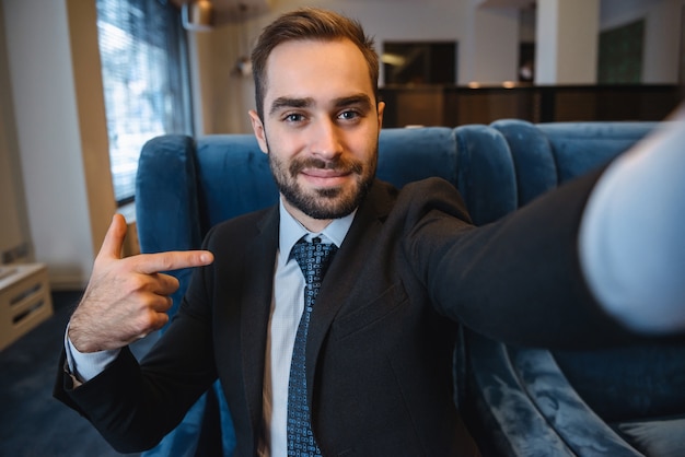 Handsome young excited businessman wearing suit sitting at the hotel lobby, using mobile phone, taking a selfie, pointing