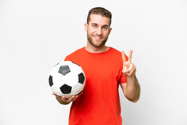 Красивый молодой футболист на изолированном белом фоне улыбается и показывает знак победы