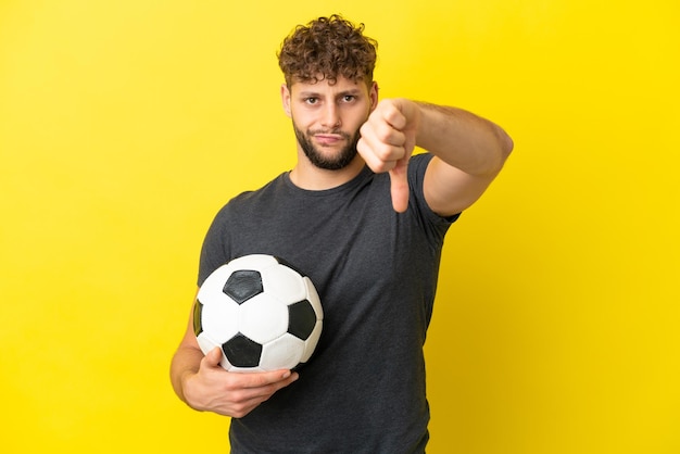 노란색 배경에 격리된 잘생긴 젊은 축구 선수 남자는 부정적인 표정으로 엄지손가락을 아래로 내립니다.