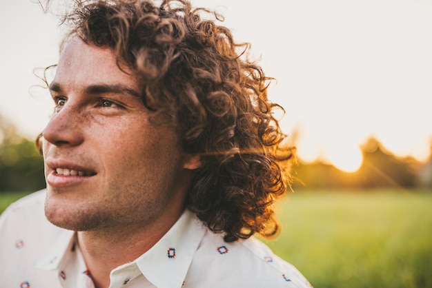 Handsome young freckled male smiling with white toothy smile and curly hair posing on the lawn in the park looking a side sunset view Copy space for your advertising People lifestyle and emotion