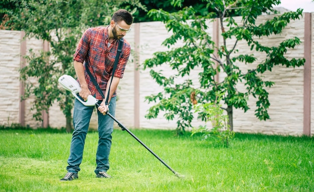 Handsome young gardener in a checkered shirt with a grass trimmer in the backyard at home outdoor