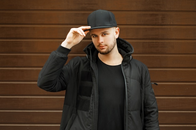 Handsome young guy in a black stylish baseball cap and black winter jacket posing near a wooden wall