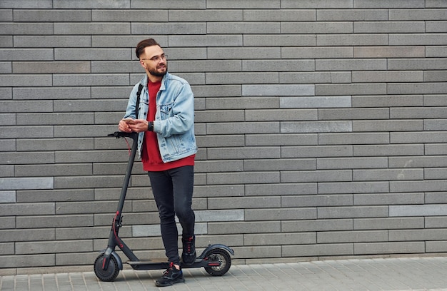 Bel giovane ragazzo in abiti casual in piedi con scooter elettrico all'aperto durante il giorno soleggiato