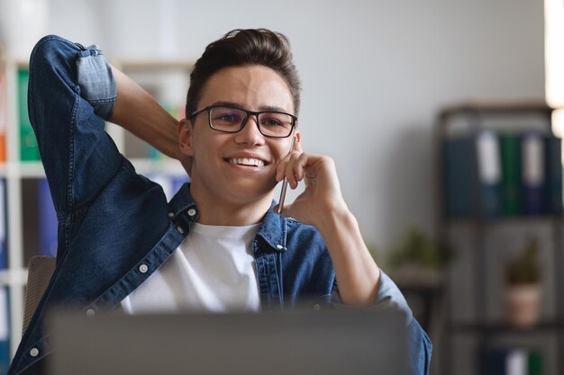 Handsome young guy in eyeglasses talking on phone at workplace in office