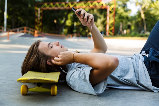Красивый молодой парень проводит время в скейт-парке, слушает музыку в наушниках, лежит на скейтборде и держит мобильный телефон