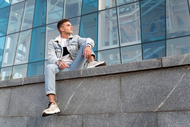 Handsome young guy in stylish clothes sits on glass facade surface. Young man in the city. Student resting after study.