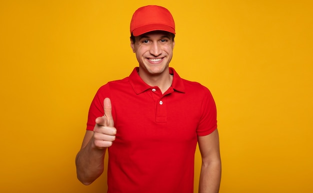 Handsome young happy courier or delivery man in red uniform is posing isolated on yellow background.