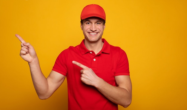 Handsome young happy courier or delivery man in red uniform is posing isolated on yellow background.