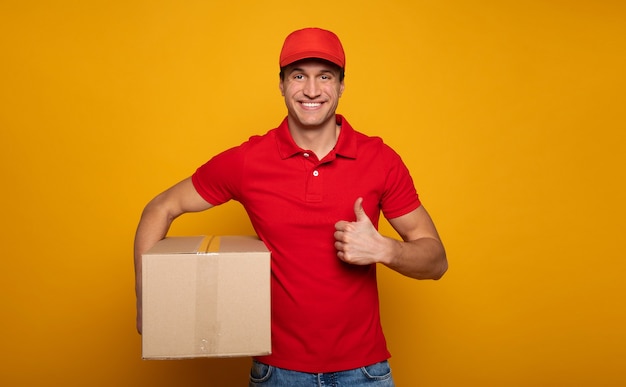 Handsome young happy courier or delivery man in red uniform is posing isolated on yellow background.