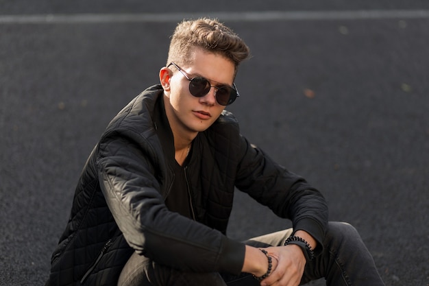 Handsome young hipster man with sunglasses in black stylish clothes sits on the street on a sunny day