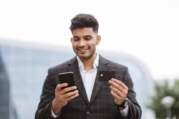 Handsome young Indian man in eyeglasses using smart phone and holding credit card