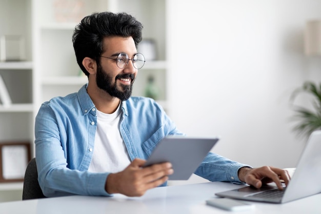 Handsome young indian man working with digital tablet and laptop at desk