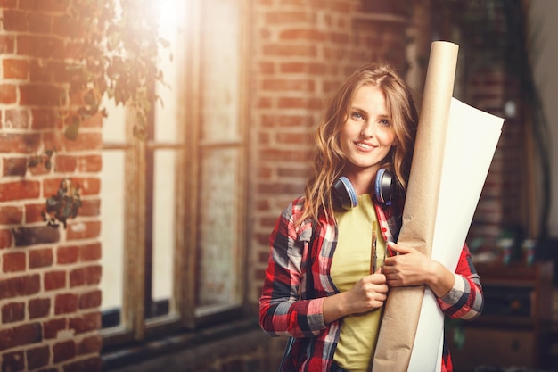 Foto giovane architetto femminile dai capelli lunghi bello che tiene i rotoli di carta e che sorride vicino alla finestra nell'ufficio moderno del sottotetto