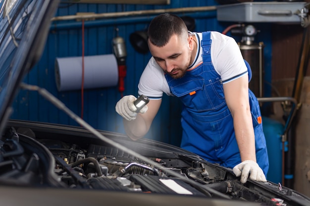 Handsome young male auto mechanic in special uniform clothes holding a flashlight, looking for breakdown and repairing under the hood in the car engine in a car workshop