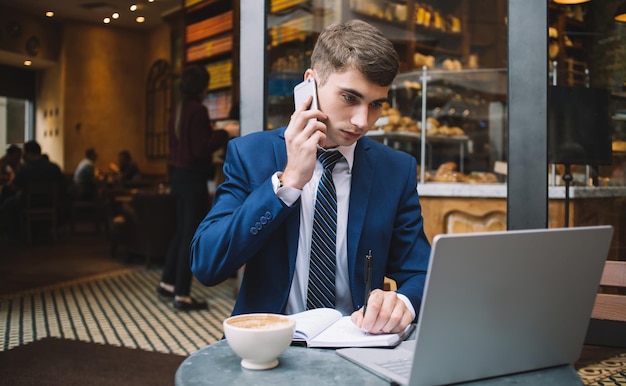 Handsome young male in elegant formal suit sitting at table in cafe making phone call while working on laptop and taking notes