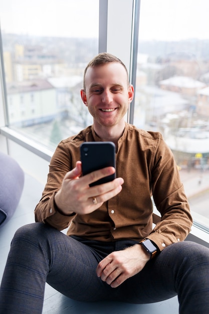 Handsome young male manager is sitting on the floor with the phone near the panoramic window. Male businessman working on a new project