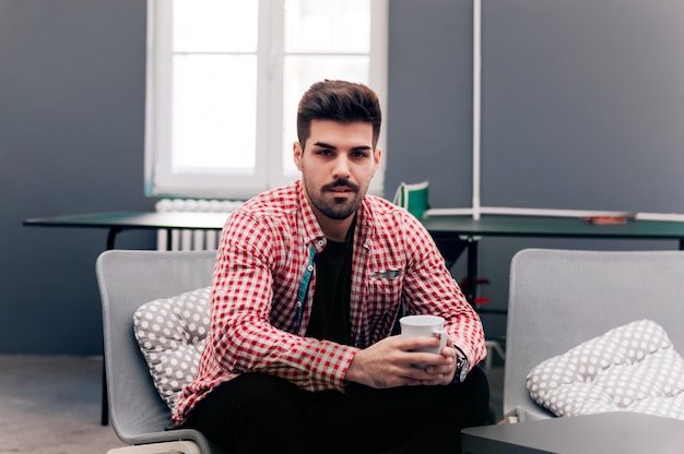 Il giovane studente maschio bello sta sedendosi nel suo ministero degli interni, tenente la tazza di caffè.