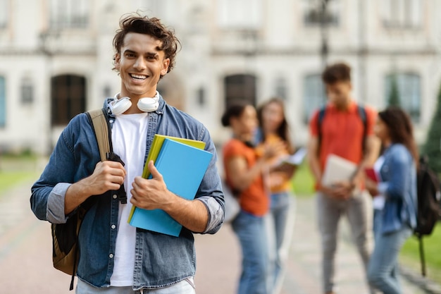 Handsome young male student with workbooks in hands posing outdoors at campus