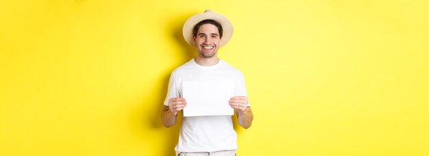 Handsome young male tourist in summer hat smiling holding blank piece of paper for your sign standin