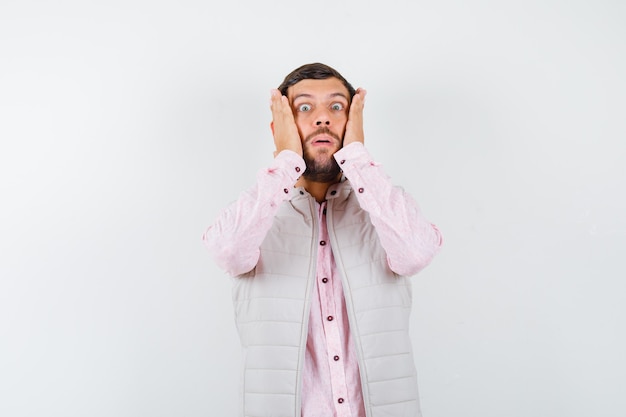 Handsome young male with hands on cheeks in shirt, vest and looking agitated. front view.