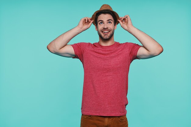 Handsome young man adjusting his fedora hat and smiling while standing against blue wall