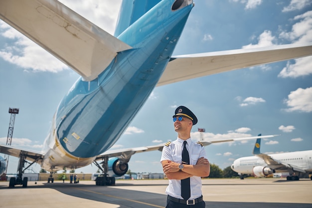 Photo handsome young man airline worker crossing arms and smiling while standing outdoors with airplanes and sky on background