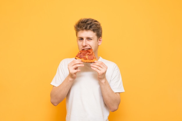 Фото Красивый молодой человек откусывает кусок аппетитной пиццы и смотрит в камеру на желтом