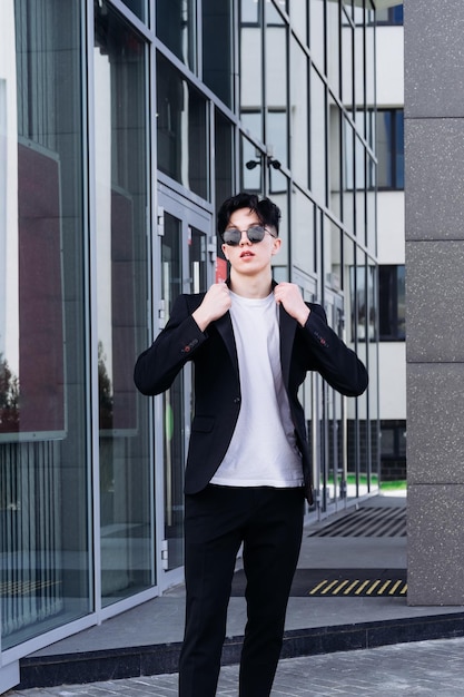 Handsome young man in a black shirt standing against the backdrop of city buildings and looking away through black sunglasses