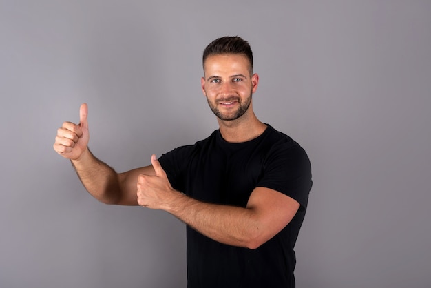 A handsome young man in a black tshirt showing thumbs up on grey