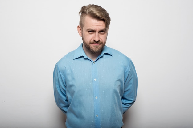 Handsome young man in a blue shirt standing at white wall with his arms crossed behind his back