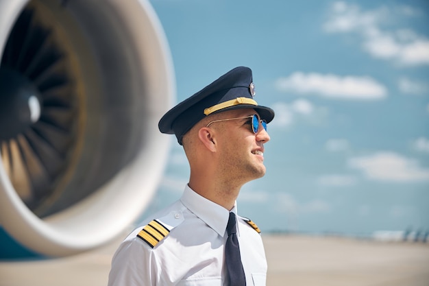 Handsome young man in captain hat looking away and smiling while standing at airport with airplane and sky on blurred background