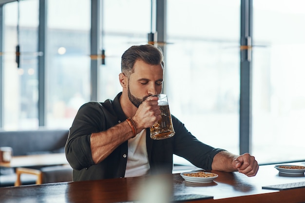 Handsome young man in casual clothing drinking beer while spending time in the pub