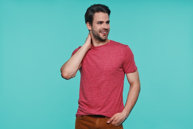 Handsome young man in casual clothing looking away and smiling while standing against blue wall