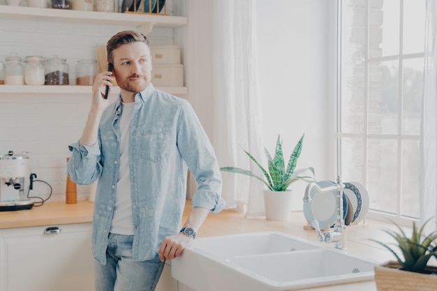 Handsome young man in casual jeans shirt speaking on cellphone in kitchen