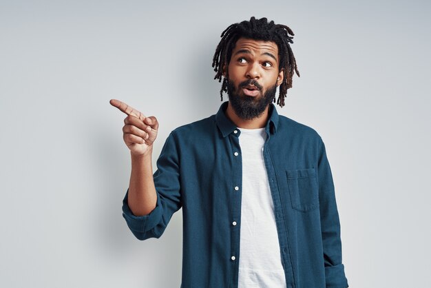 Handsome young man in casual wear pointing copy space while standing against grey wall