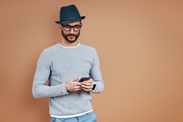 Handsome young man in casual wear using smart phone and looking at camera while standing against brown wall