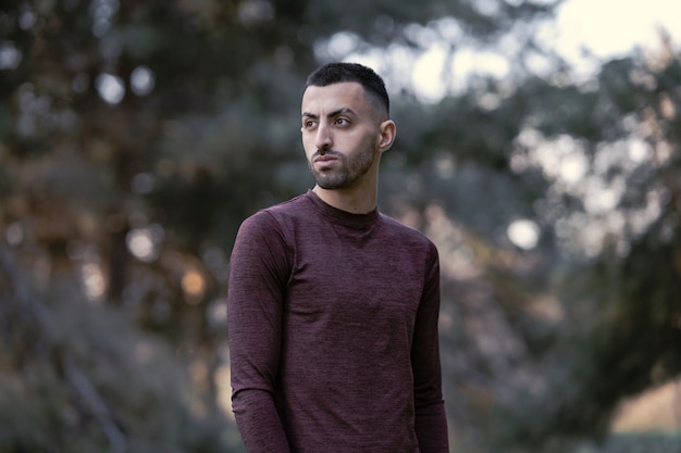 Photo a handsome young man dressed in casual clothes and a cap stands in a thick coniferous forest and looks at the cameras with a serious face.