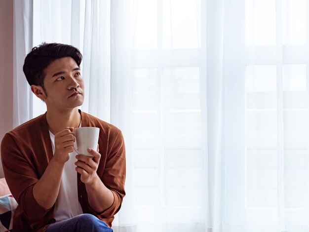 Handsome young man drinking at home next to window curtains