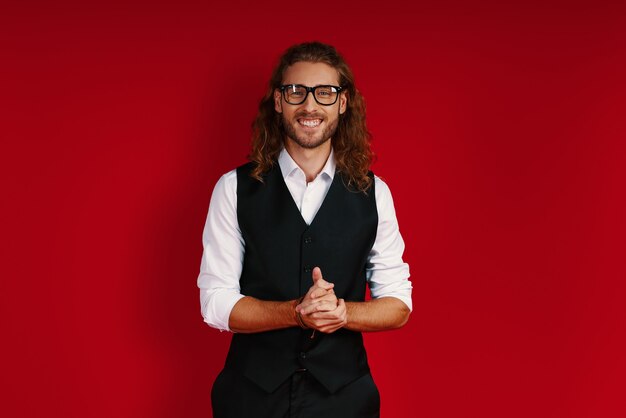 Handsome young man in elegant clothing looking at camera and smiling while standing against red wall