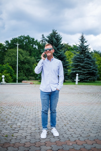 A handsome young man of European appearance wearing sunglasses is dressed in a shirt and jeans. The guy walks down the street, he is stylishly dressed