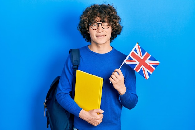 セクシーな表情、陽気で幸せそうな顔でカメラを見てまばたき英国旗を保持しているハンサムな若い男の交換留学生。