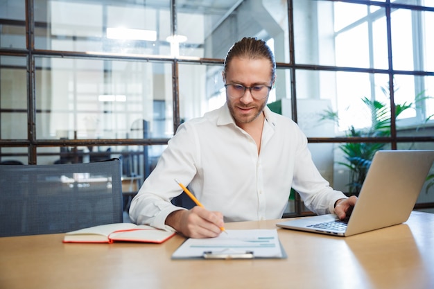 handsome young man in eyeglasses making notes and working with laptop while sitting at table