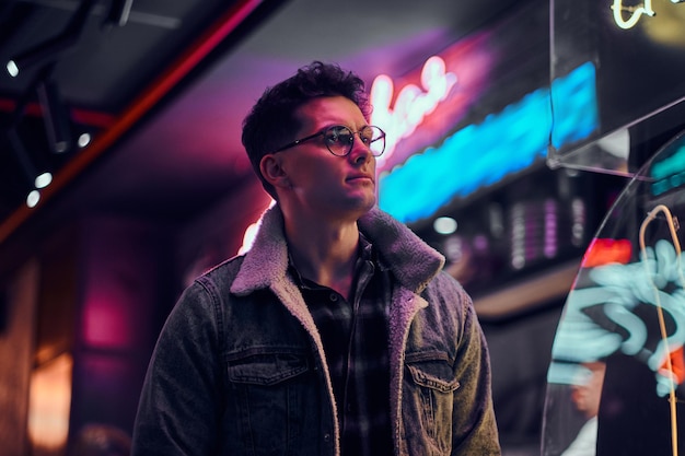 A handsome young man fashionably dressed standing in the street at night. Illuminated signboards, neon, lights.